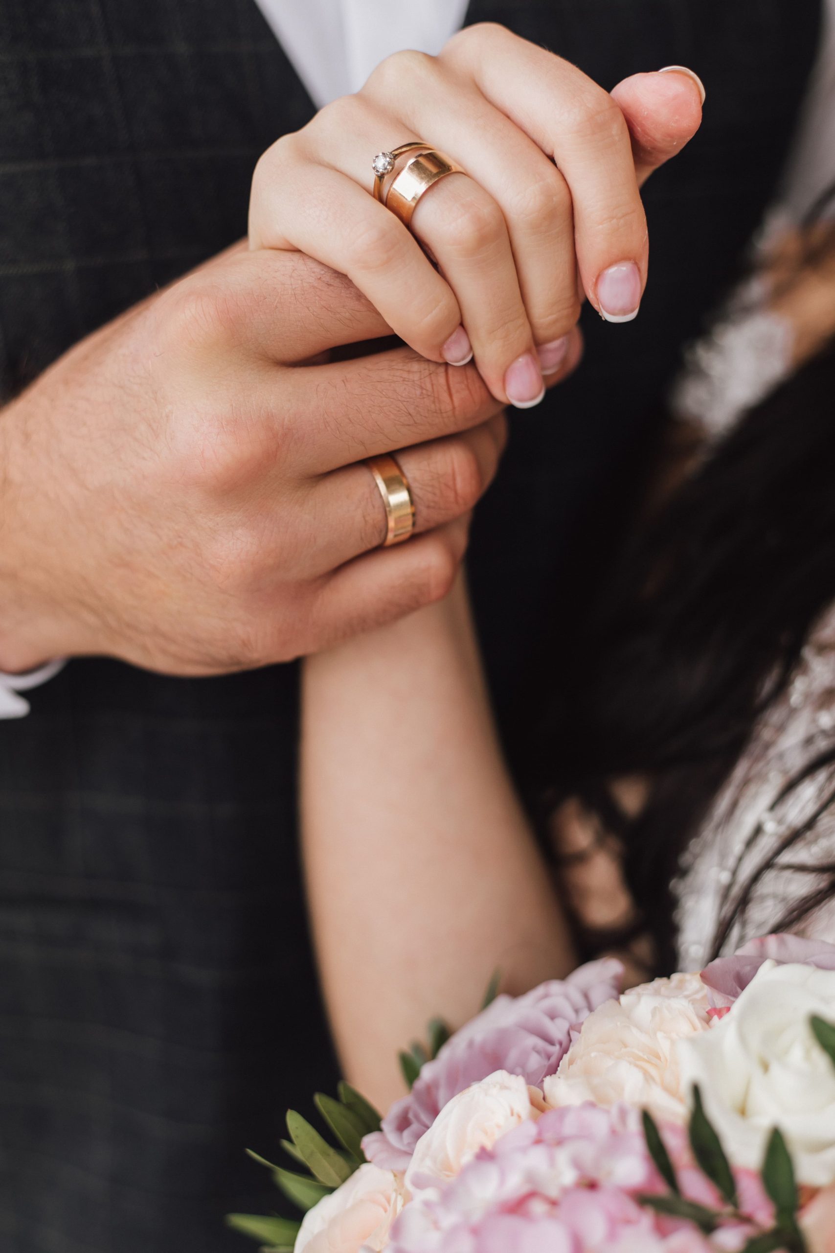 mani-di-una-moglie-e-un-marito-con-anelli-di-fidanzamento-e-matrimonio-e-parte-del-bouquet-floreale (1)